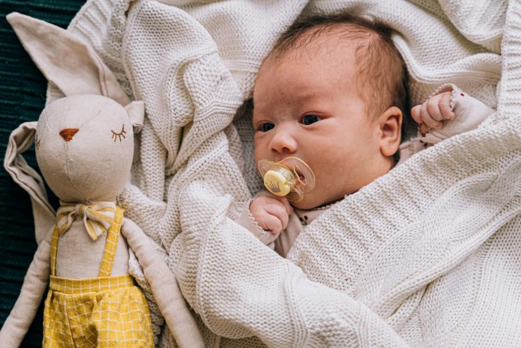 Newborn baby with a pacifier lying on a white knitted blanket next to a soft toy bunny in a yellow outfit. The baby looks calm and comfortable, representing the use of baby health products for soothing and care.