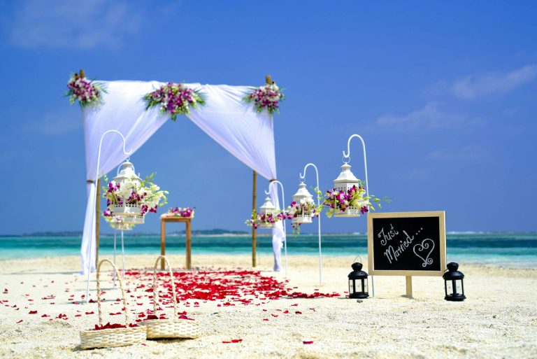 Beach destination wedding setup with a beautiful floral arch, lanterns, and a 'Just Married' sign. Ideal wedding registry gifts for a destination wedding include travel-friendly items and cash funds.