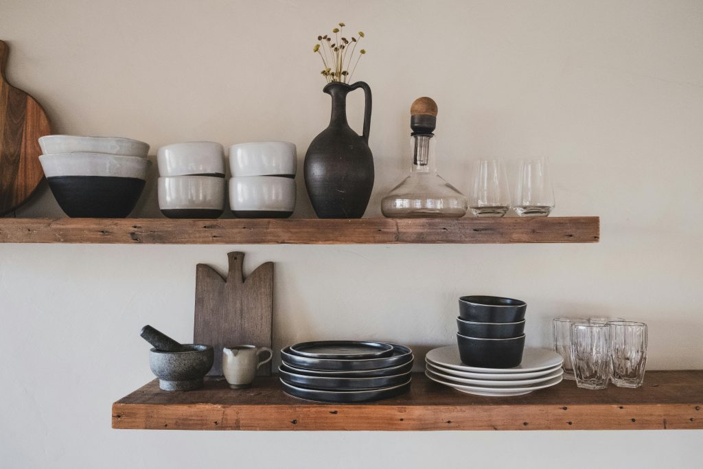 Wedding registry must-haves for the culinary duo: Rustic wooden shelves displaying a variety of kitchen essentials, including ceramic bowls, plates, a decanter, a mortar and pestle, and elegant glassware, perfect for a couple passionate about cooking.