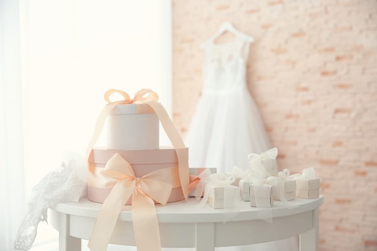 Elegant wedding gifts under $100 displayed on a white table with pastel pink ribbons, featuring a bridal dress in the background. Discover the best affordable wedding gift ideas to impress the newlyweds.