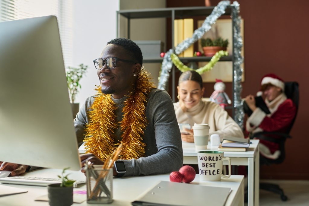 Young smiling businessman with xmas decoration on neck analyzing online data on computer screen against female colleague with smartphone
