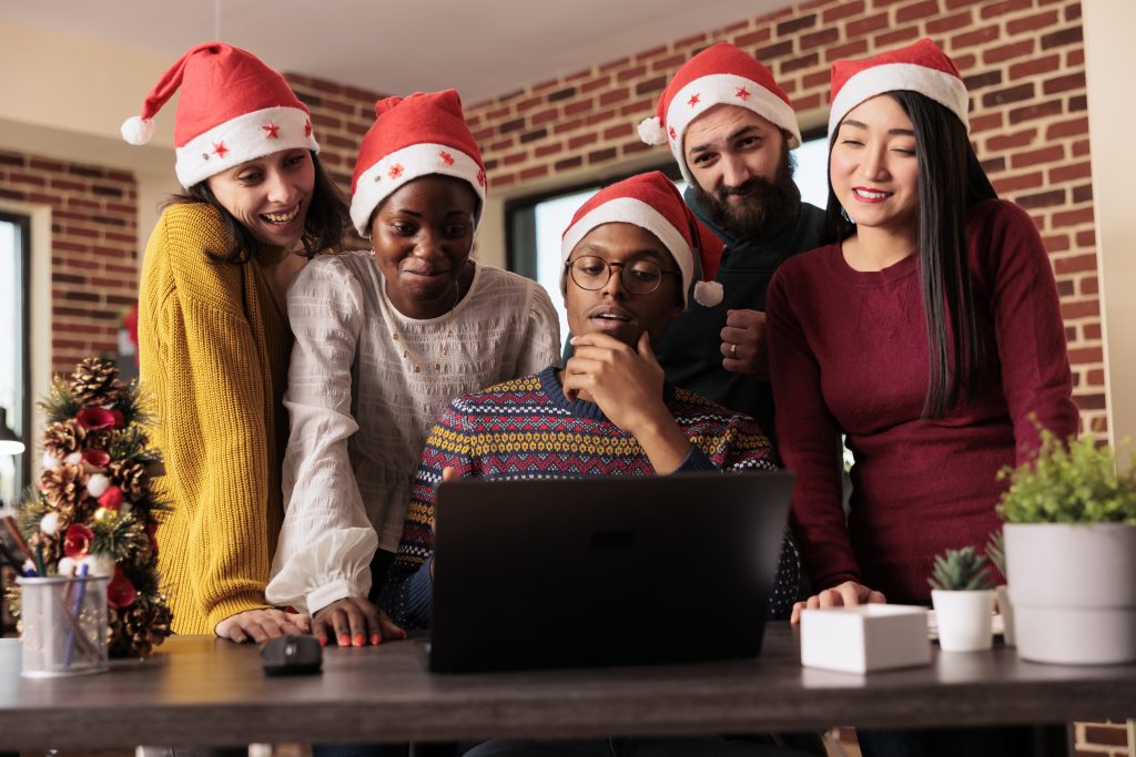Cheerful colleagues checking sales annual report results on laptop before celebrating christmas in office. Smiling happy coworkers working for project deadline together in festive decorated workplace