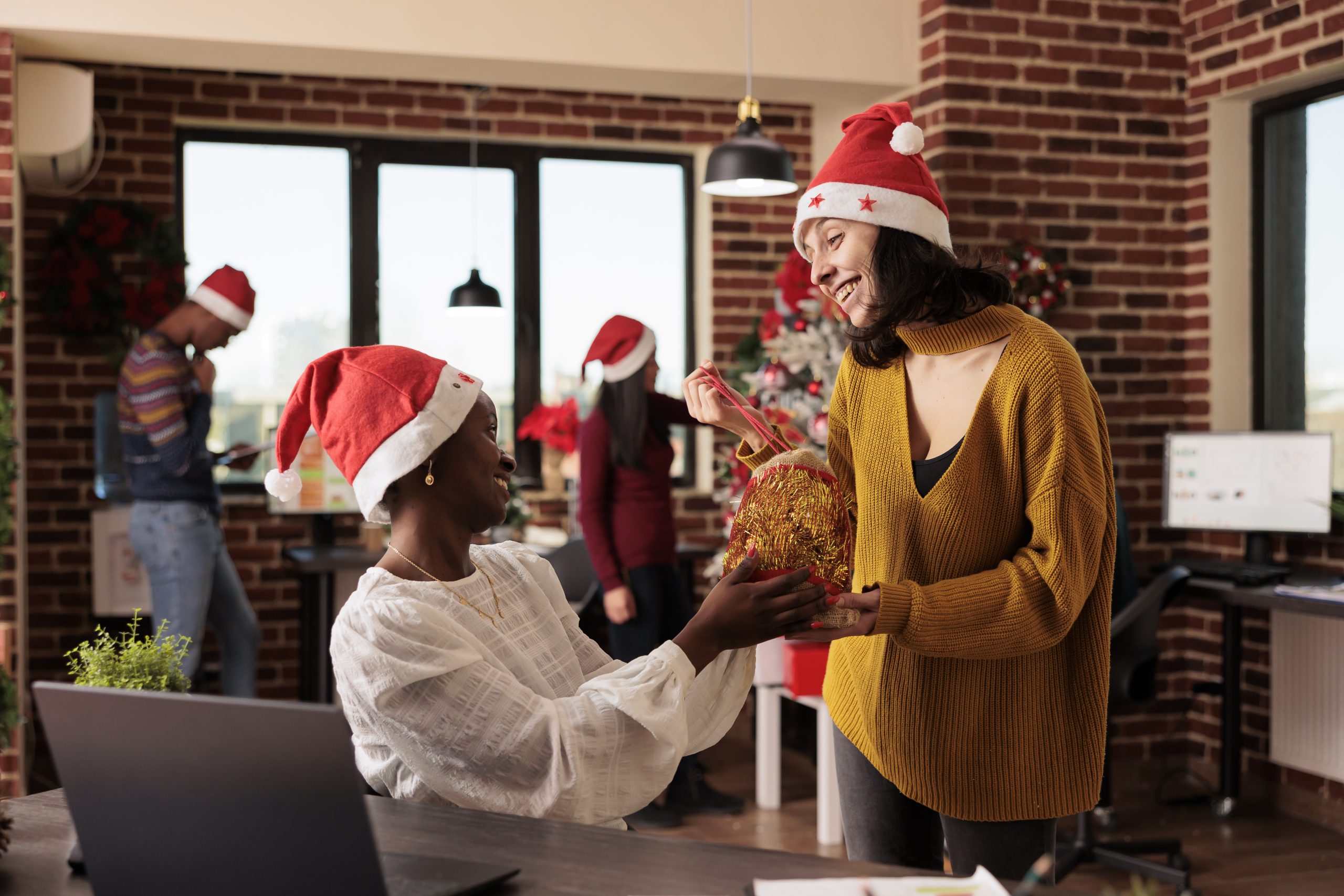 Smiling diverse women colleagues in santa hats exchanging christmas presents at startup company workplace. Cheerful caucasian employee sharing festive gift bag to coworker in decorated office