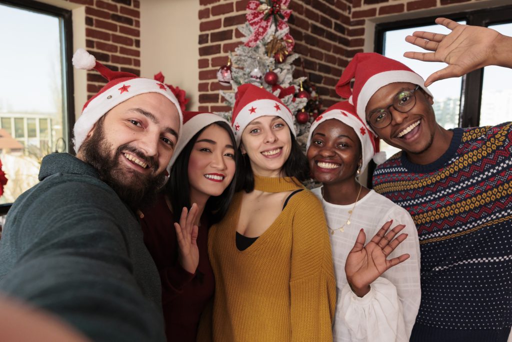 Diverse colleagues taking photos in festive office, celebrating xmas holiday with christmas tree and decorations. People taking pictures with seasonal ornaments in business space.