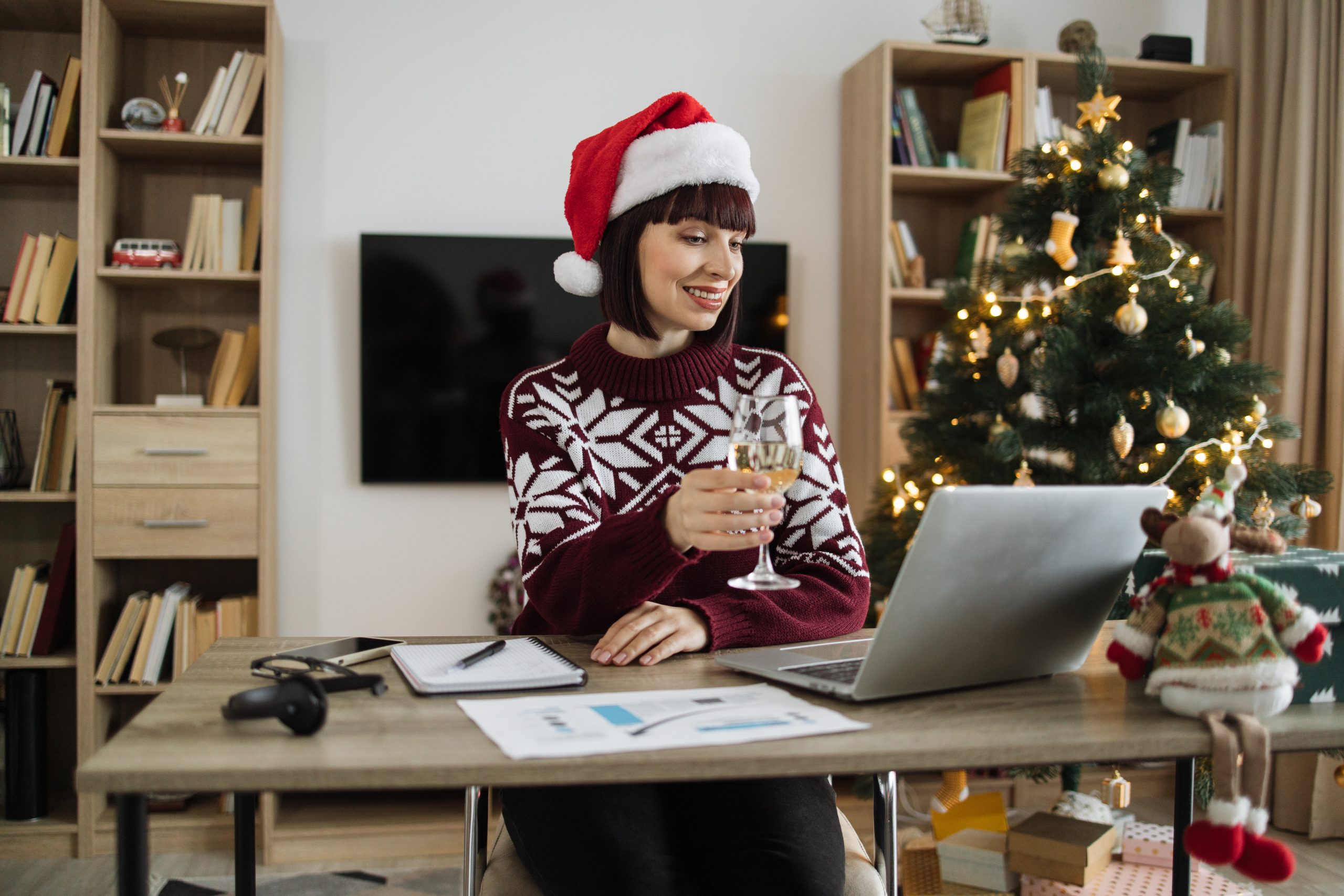 Happy caucasian female professional manager in Santa hat holding glass with champagne while talking to client over video call while staying at home on background of Christmas tree.