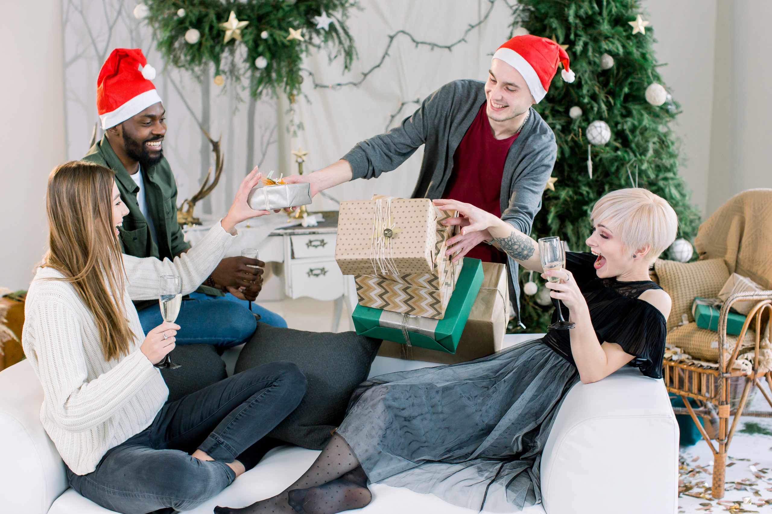 Group of happy people having a great time, celebrating New Year. Two girls and african boy sitting on white sofa and drinking champagne, and white boy behind is giving Christmas presents for them