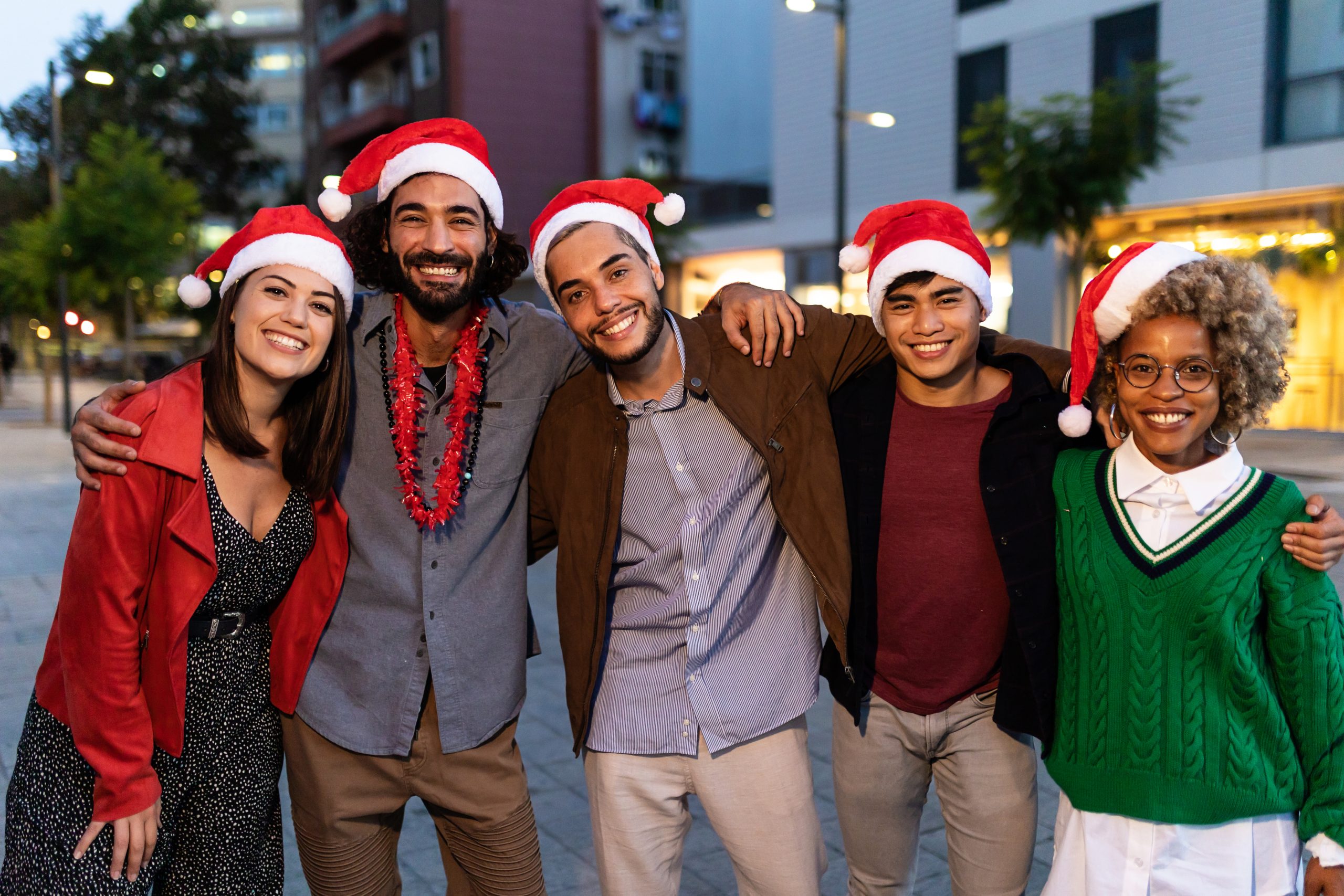 Group portrait of multiracial friends wearing Santa Claus hat standing outdoors and hugging each other at christmas day - Celebration and xmas holidays concept