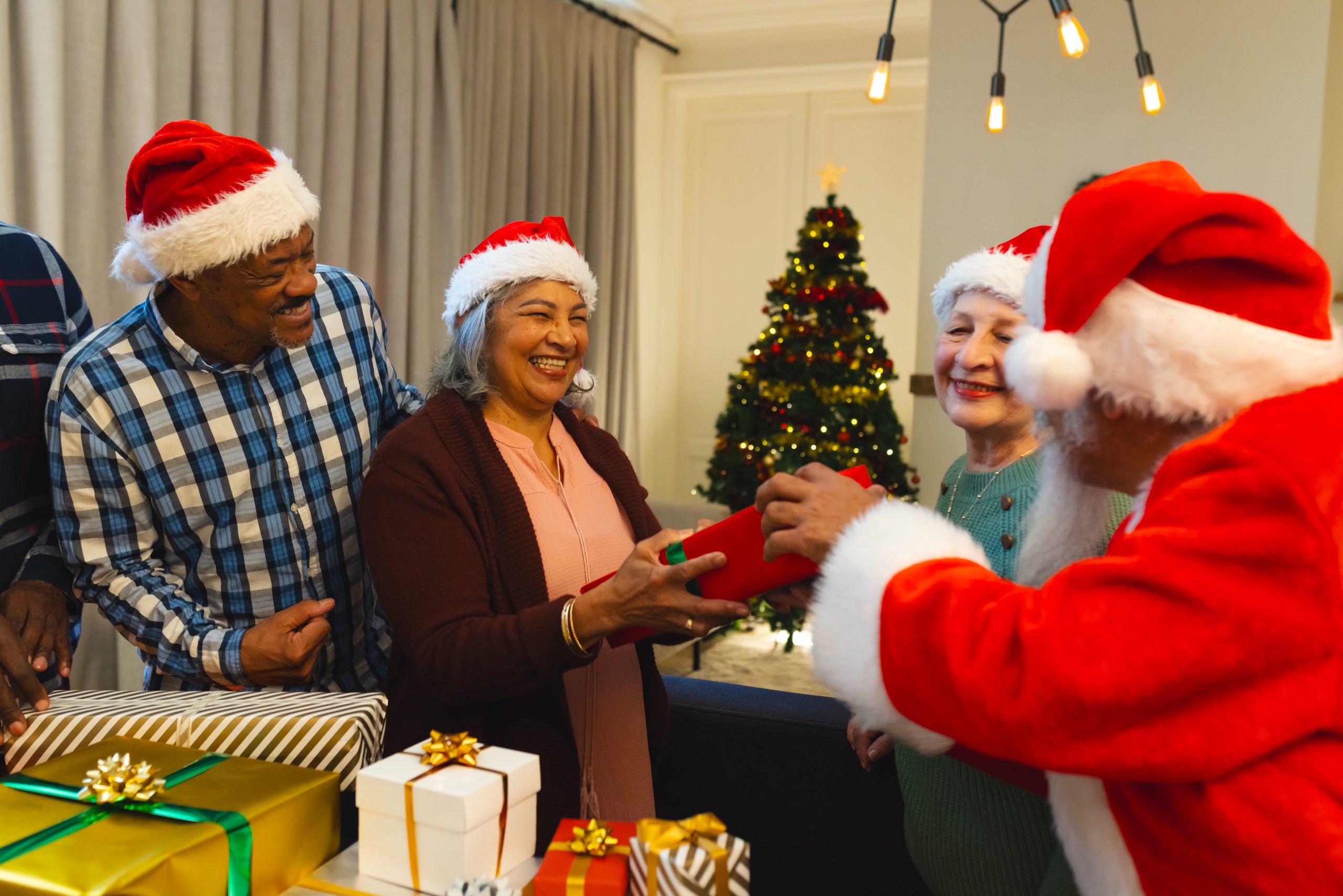 Happy biracial man in santa clothes giving gifts to friends in living room at home. Retirement, christmas, celebration, friendship, togetherness and senior lifestyle, unaltered.