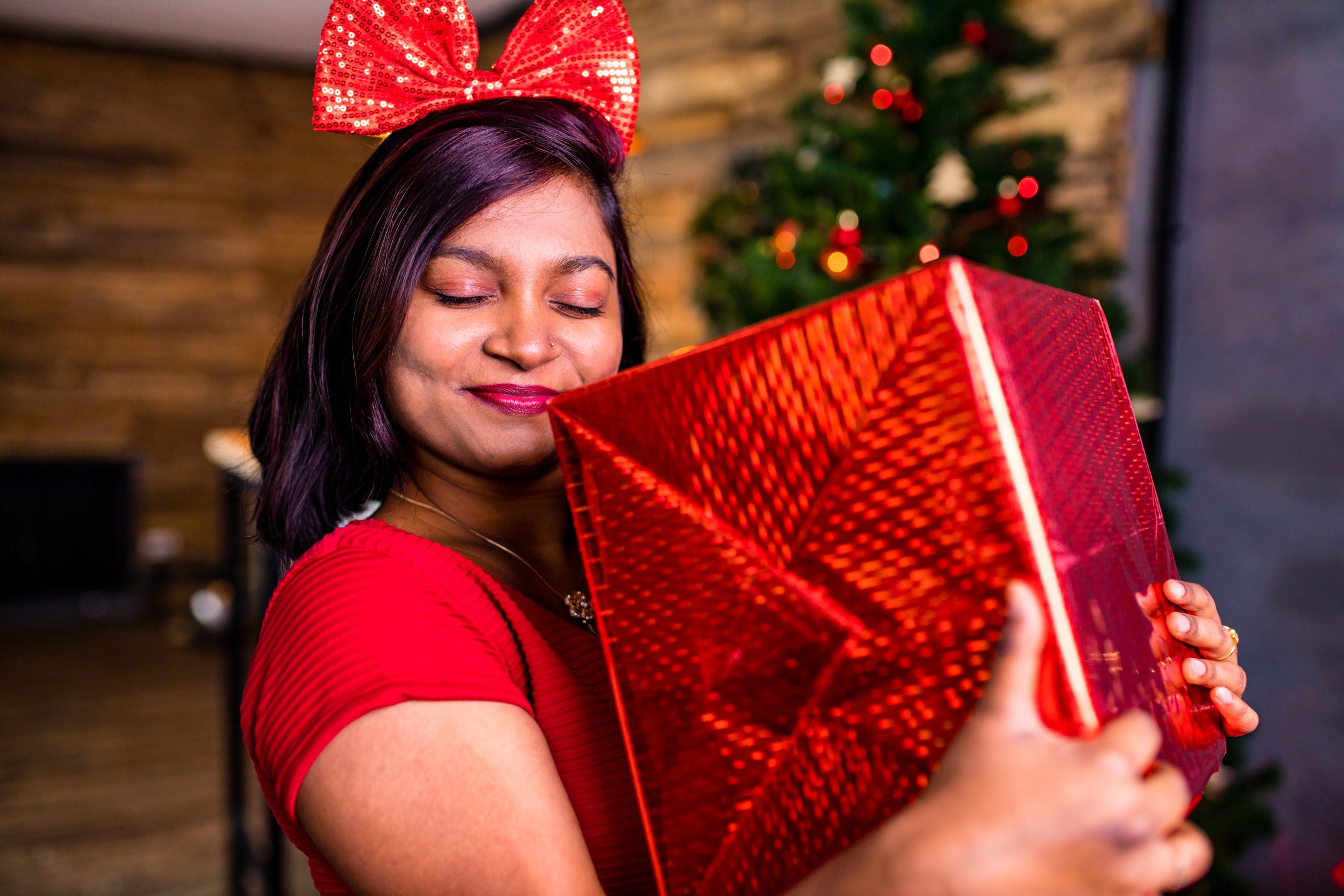 indian woman in red dress holding big box with bowl in living room spruce Christmas tree lights garland background .