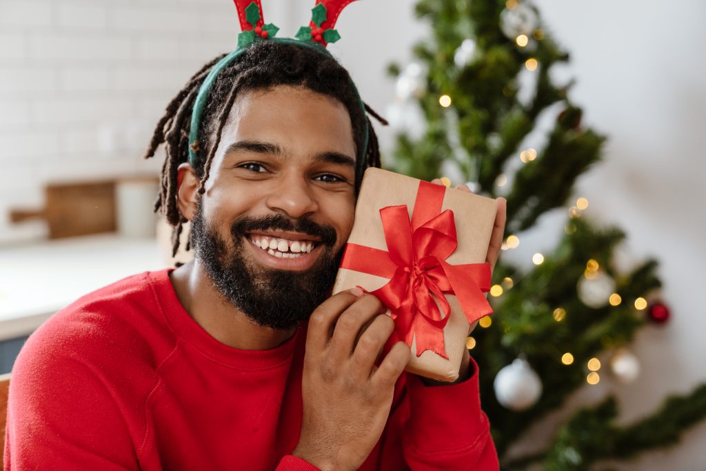 Joyful african american man in toy deer horns smiling and holding gift box in cozy room