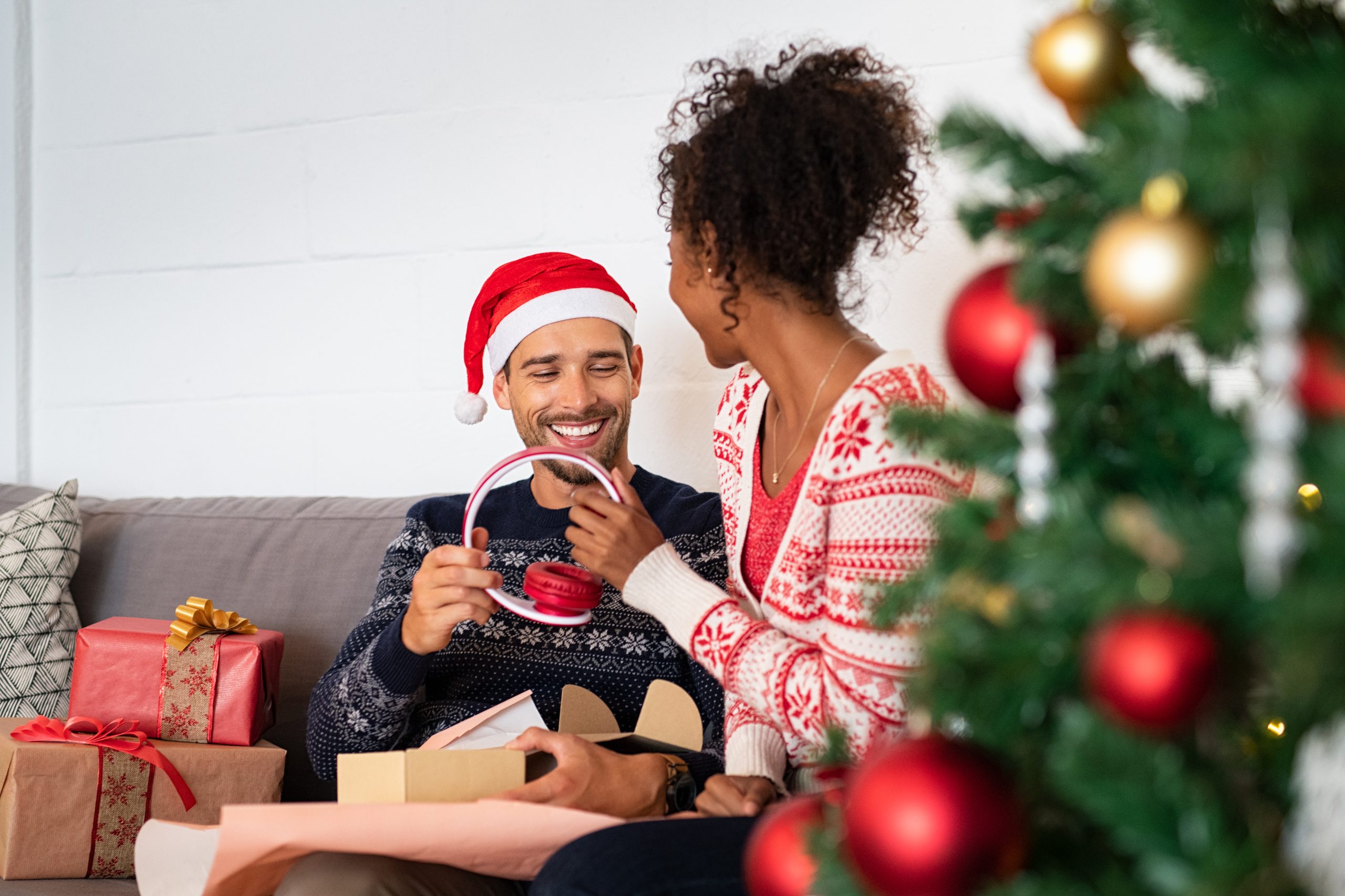 Happy man receiving red wireless headphone as christmas present. Multiethnic couple unwrapping xmas present on sofa holding new earphone. Husband wearing santa claus hat and wife opening xmas present with headphone in it.