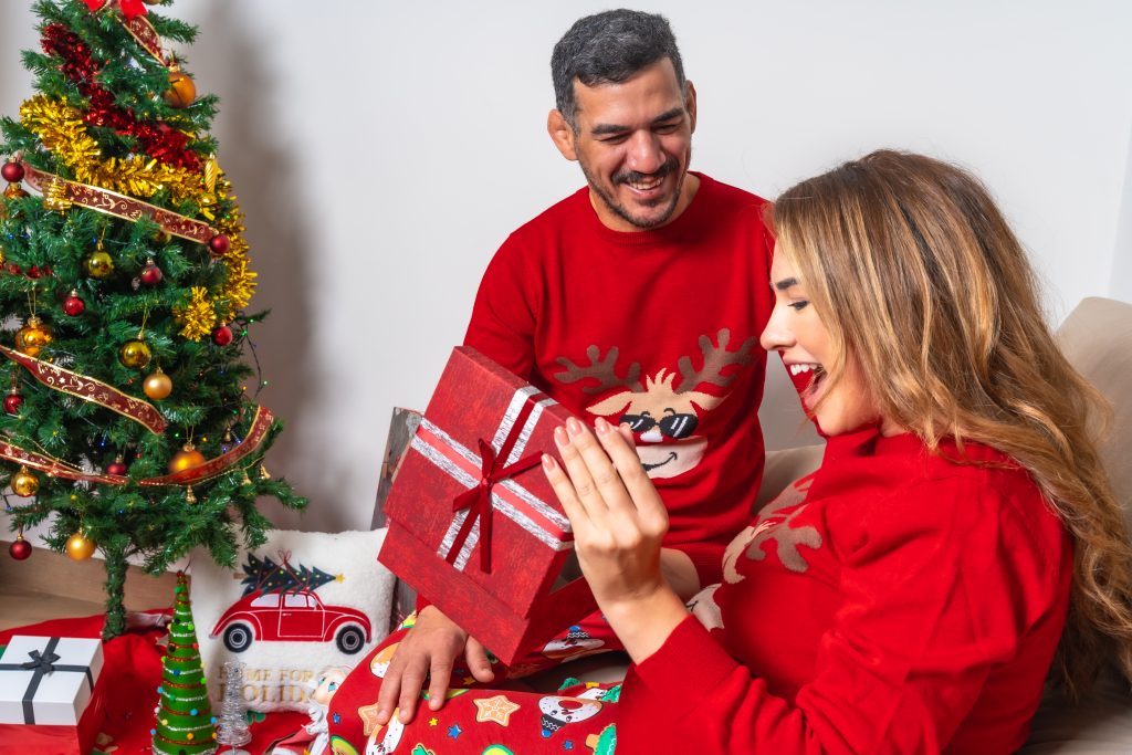 Young couple sitting on the sofa with decoration and red Christmas clothes, boyfriend giving a gift to the girlfriend and very happy girlfriend