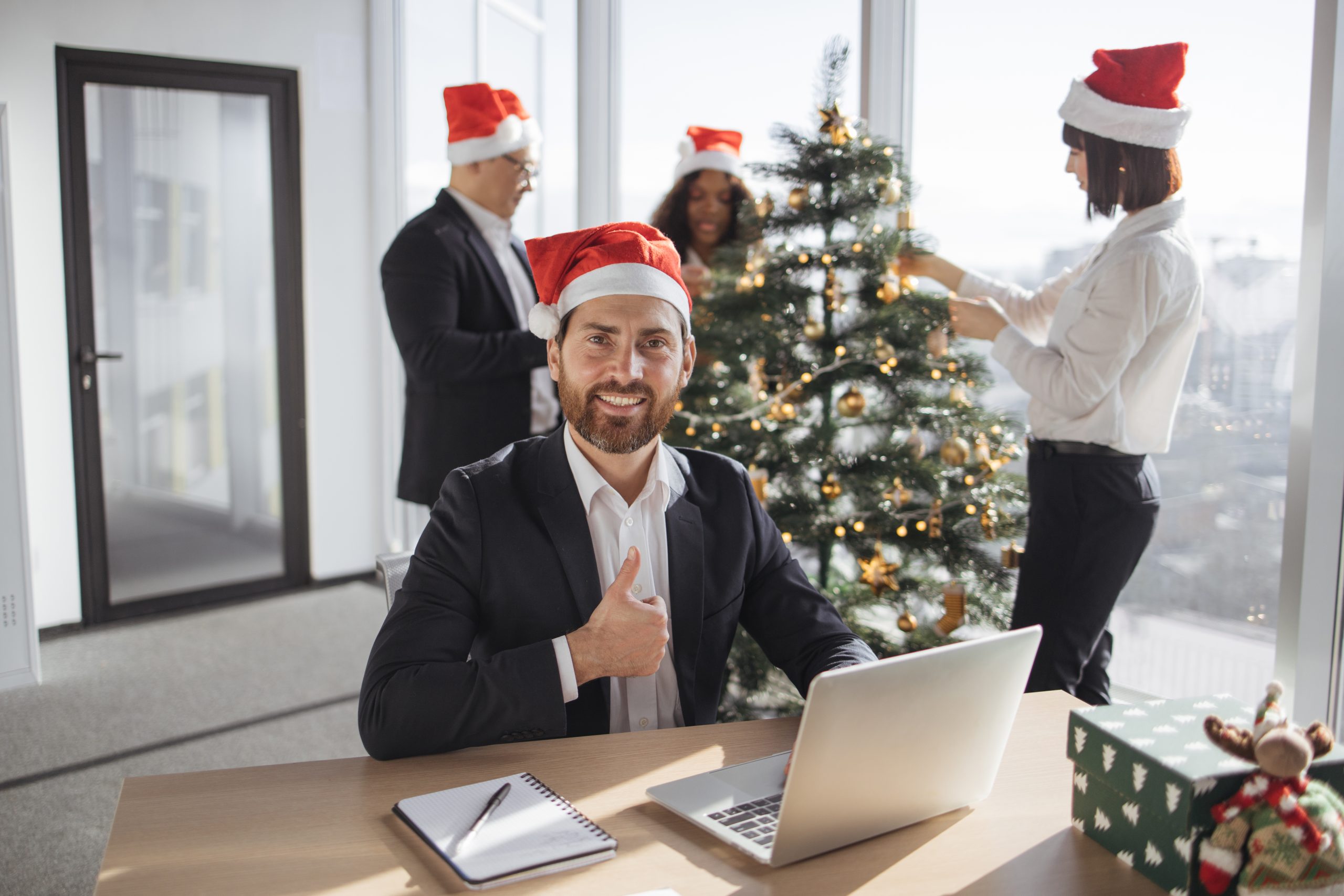 Attractive Caucasian businessman employee in suit and santa hat sitting at desk using laptop in christmas decorated office. Young corporate worker in festive workplace looking at camera.