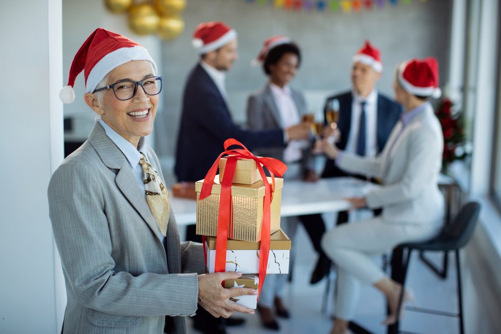 Happy mature businesswoman with gift boxes coming to Christmas party in the office and looking at camera. Her colleagues are celebrating in the background.