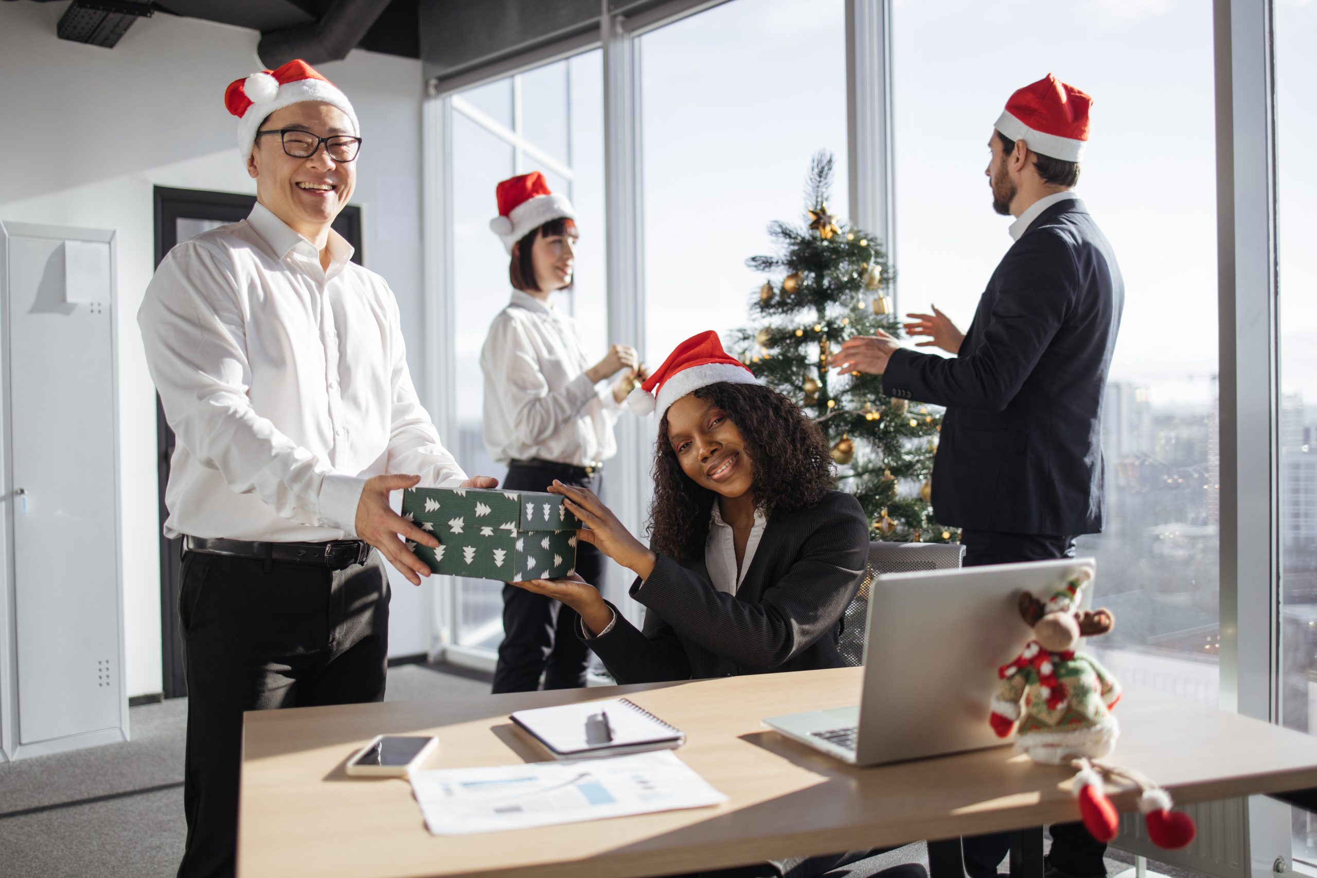 Smiling diverse colleagues exchanging christmas gifts at corporate party in decorated festive office. Company employee giving present to coworker while celebrating winter holiday at workplace.