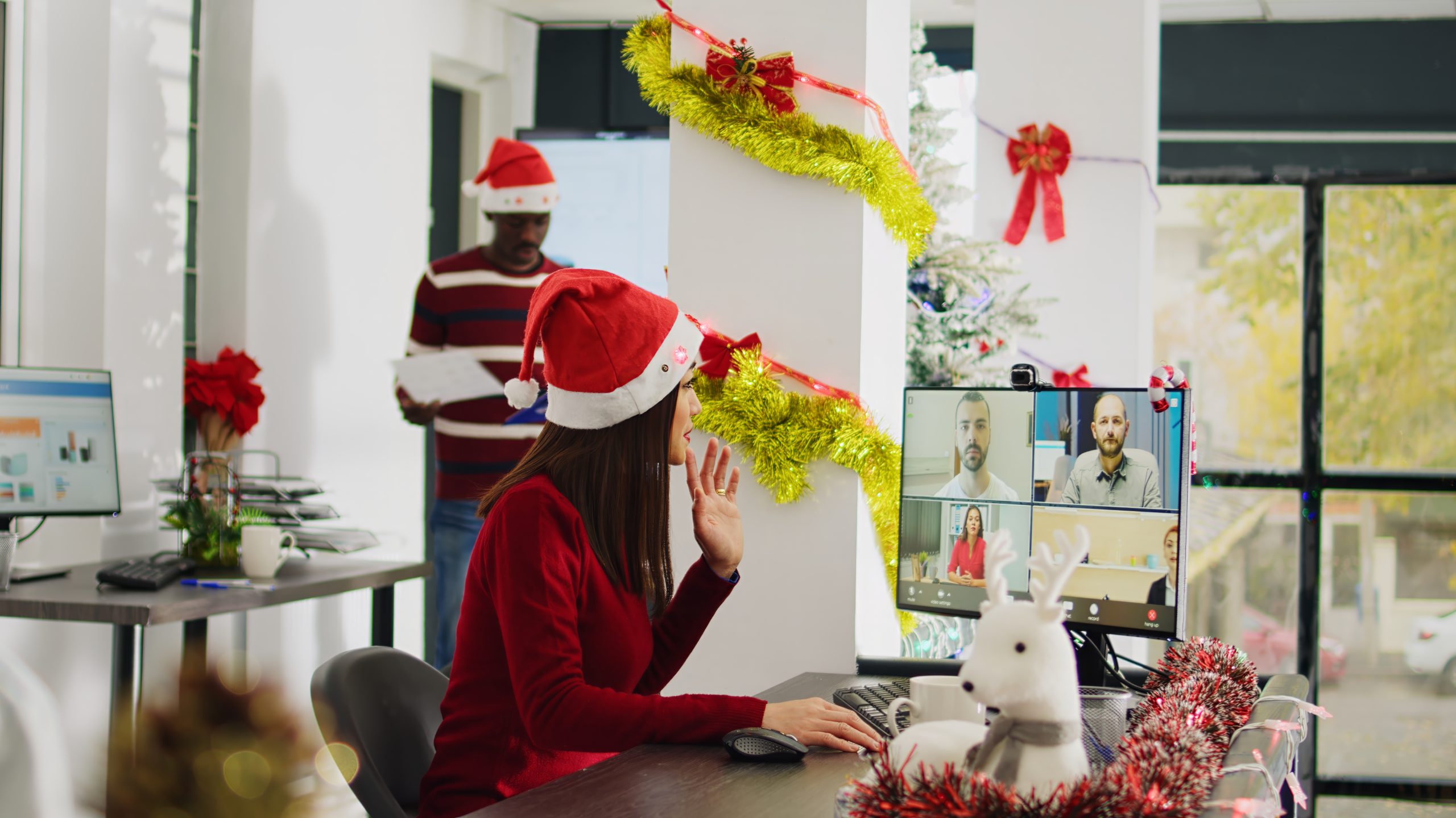 Asian worker in online videocall with colleagues during Christmas season in festive decorated office. Employee using computer webcam for remote teleconference with team in busy xmas ornate workplace