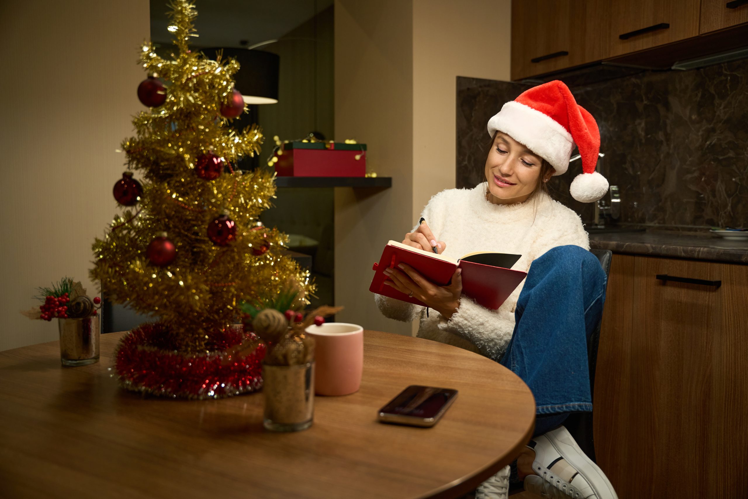 Young thoughtful and smiling beautiful caucasian woman wearing santa hat writing her notes and wishes in notebook at table during Christmas or New Year at home at night. Winter holidays celebrating