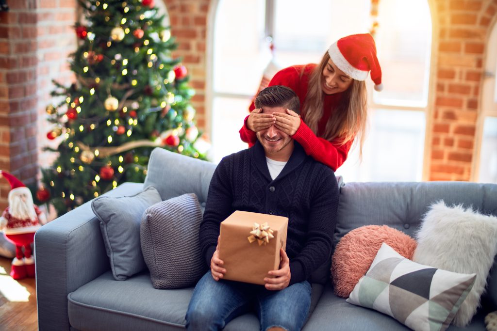 man holding Christmas gift while his girlfriend covers his eyes from behind