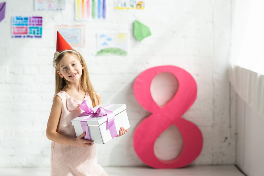 adorable smiling kid in party cap holding present and posing near decorative pink number 8
