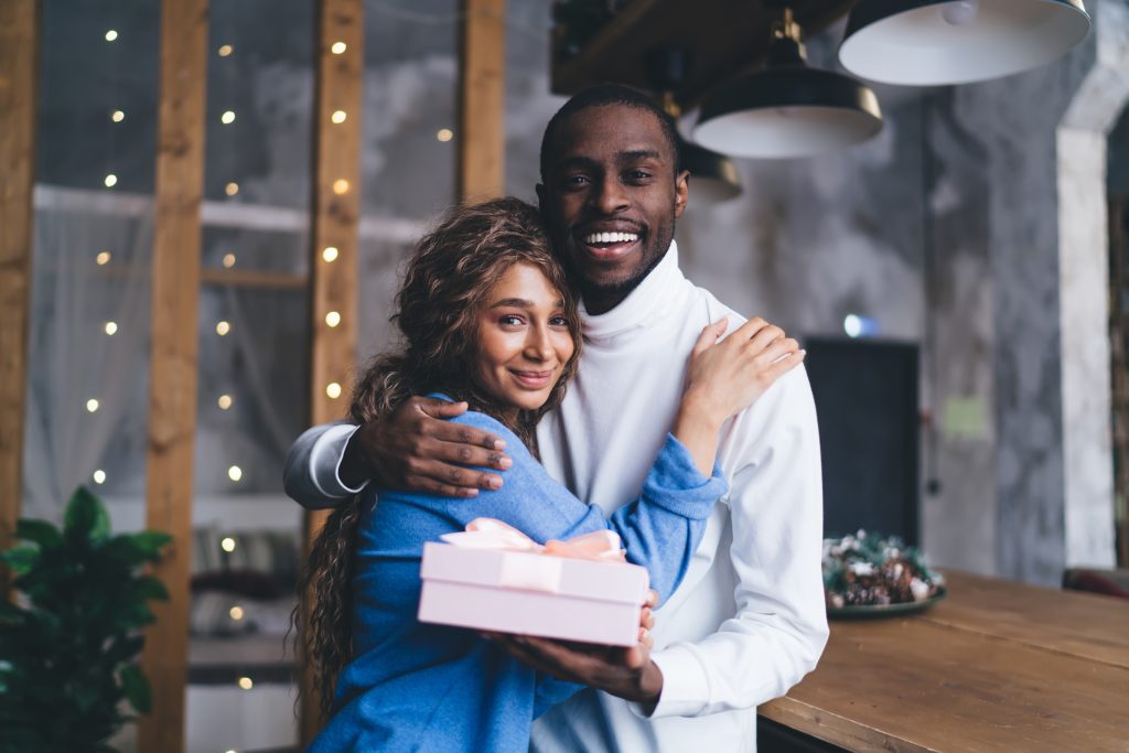 joyful Black couple embraces in cozy indoor , husband man presenting pink gift box. Woman smile, reflecting heartwarming surprise. soft-focus lights, suggesting festive or romantic occasion
