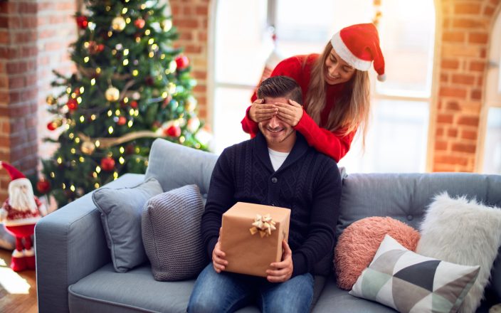 man holding Christmas gift while his girlfriend covers his eyes from behind