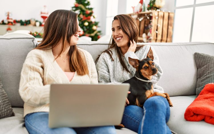 Two women using laptop sitting with dog by christmas tree at home