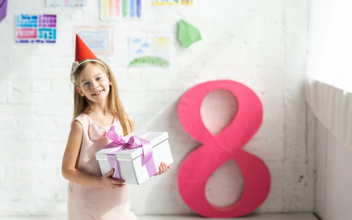 adorable smiling kid in party cap holding present and posing near decorative pink number 8