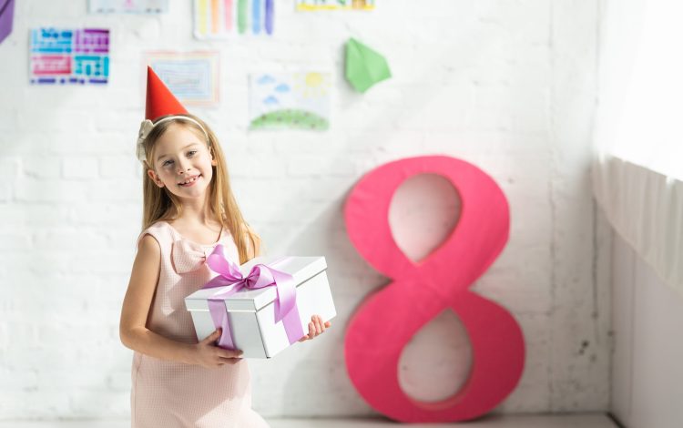 adorable smiling kid in party cap holding present and posing near decorative pink number 8