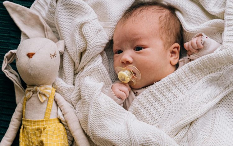 Newborn baby with a pacifier lying on a white knitted blanket next to a soft toy bunny in a yellow outfit. The baby looks calm and comfortable, representing the use of baby health products for soothing and care.