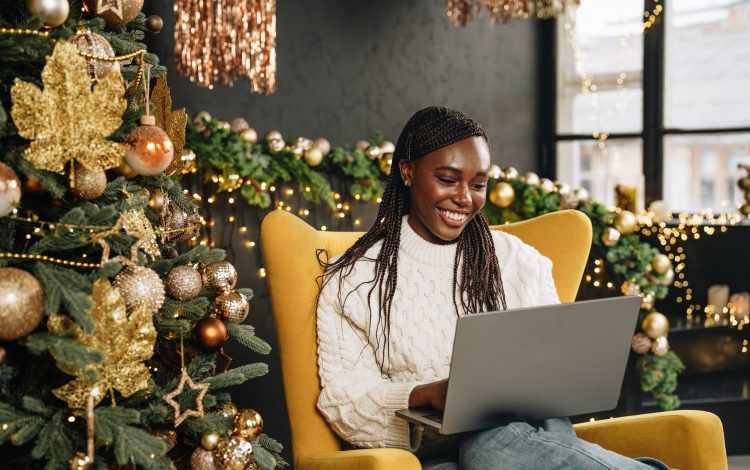 Beautiful young african female sitting with laptop near Christmas tree at home close up