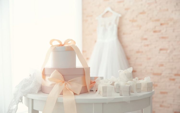 Elegant wedding gifts under $100 displayed on a white table with pastel pink ribbons, featuring a bridal dress in the background. Discover the best affordable wedding gift ideas to impress the newlyweds.