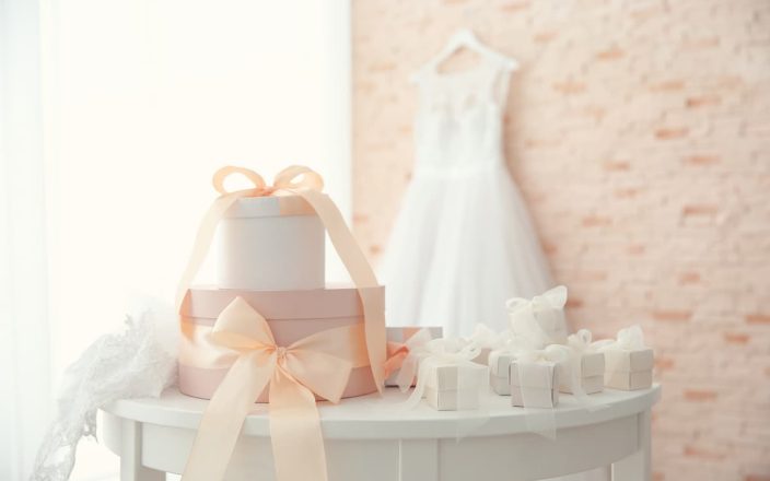 Elegant wedding gifts under $100 displayed on a white table with pastel pink ribbons, featuring a bridal dress in the background. Discover the best affordable wedding gift ideas to impress the newlyweds.