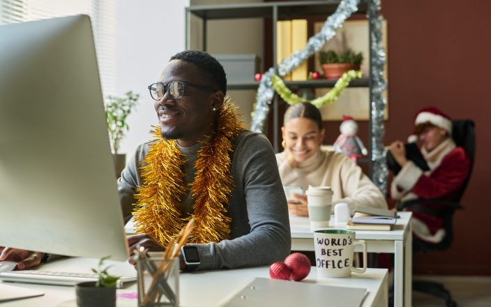 Young smiling businessman with xmas decoration on neck analyzing online data on computer screen against female colleague with smartphone