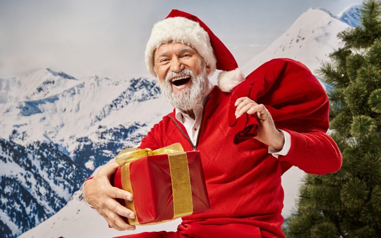 cheerful man dressed as Santa smiling happily at camera holding gift bag and present, winter