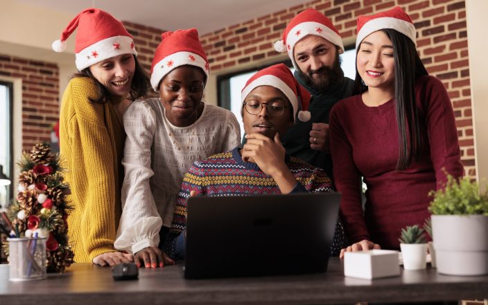 Cheerful colleagues checking sales annual report results on laptop before celebrating christmas in office. Smiling happy coworkers working for project deadline together in festive decorated workplace