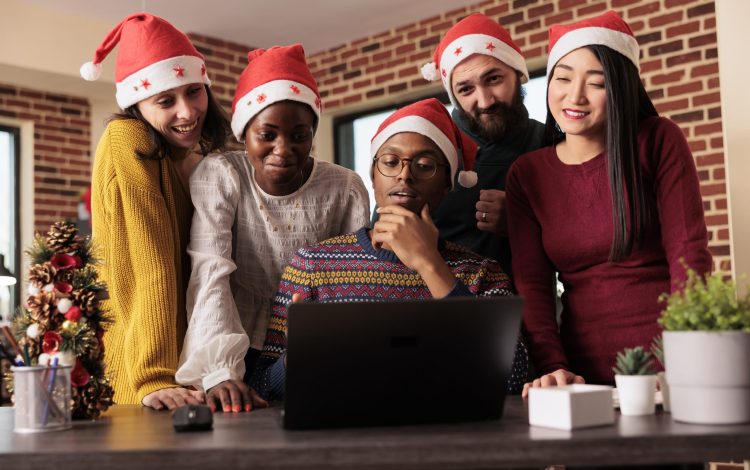 Cheerful colleagues checking sales annual report results on laptop before celebrating christmas in office. Smiling happy coworkers working for project deadline together in festive decorated workplace