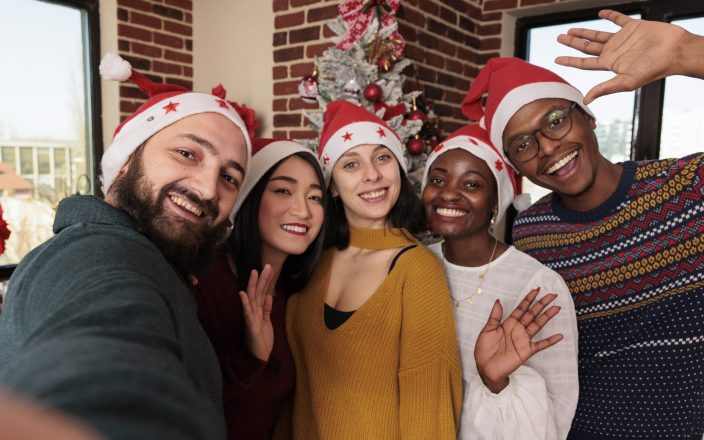 Diverse colleagues taking photos in festive office, celebrating xmas holiday with christmas tree and decorations. People taking pictures with seasonal ornaments in business space.