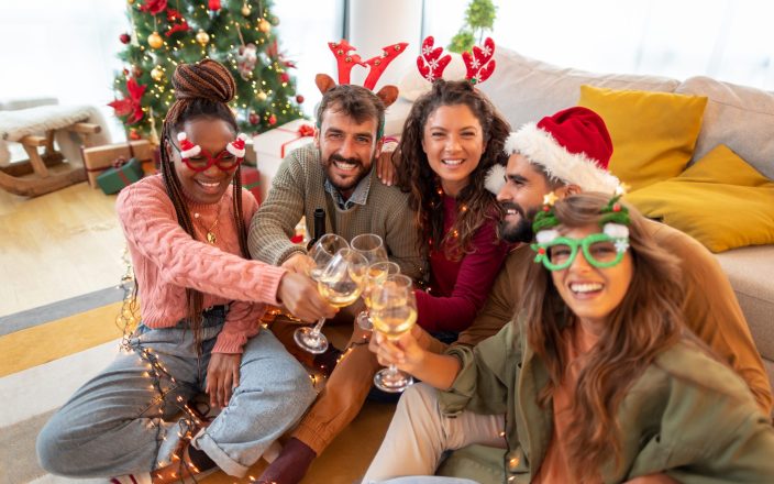 Cheerful young friends sitting by nicely decorated Christmas tree, celebrating Christmas at home, making a toast and drinking wine