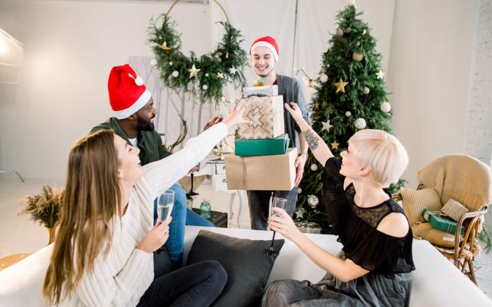 Picture showing group of friends with Christmas presents, celebrating Christmas or New Year in cozy decorated living room. Two boys, African and Caucasian in santa hats with two Caucasian girls