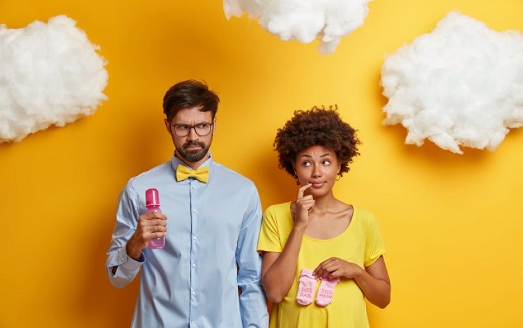 Husband and wife await for baby, pose with feeding bottle and newborn booties, think over name of future child, get ready to become parents, isolated on yellow background, white clouds overhead