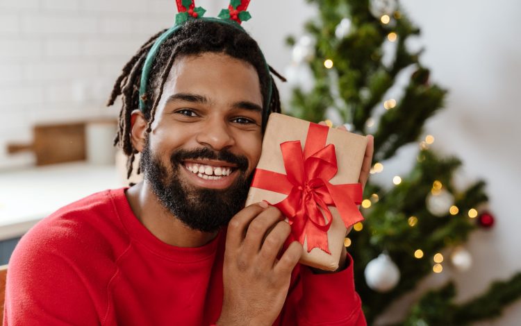 Joyful african american man in toy deer horns smiling and holding gift box in cozy room