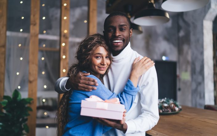 joyful Black couple embraces in cozy indoor , husband man presenting pink gift box. Woman smile, reflecting heartwarming surprise. soft-focus lights, suggesting festive or romantic occasion
