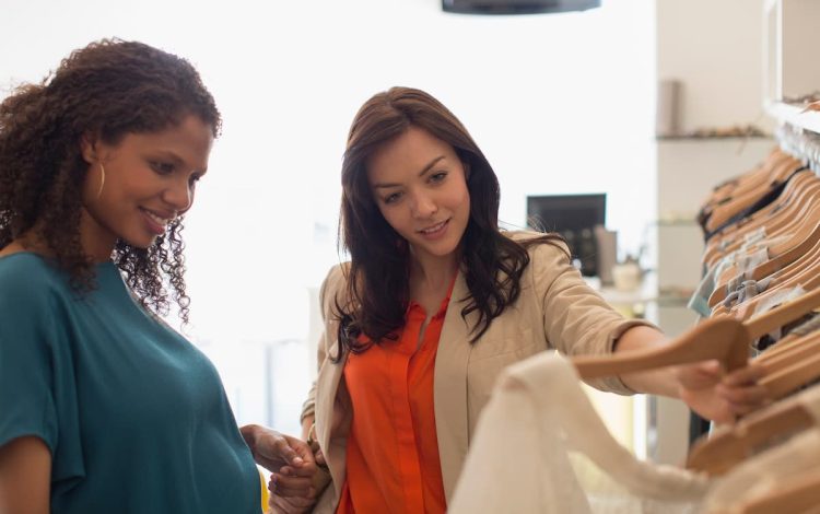 Two women shopping for maternity clothes in a stylish boutique, with one holding up a dress and both smiling, showcasing options for a chic maternity wardrobe. Keywords: maternity wardrobe, baby registry.