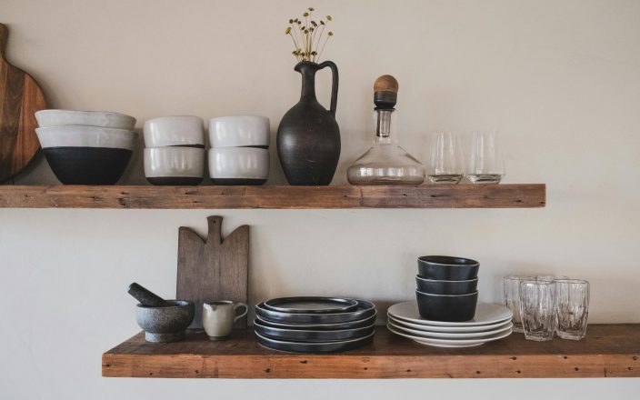 Wedding registry must-haves for the culinary duo: Rustic wooden shelves displaying a variety of kitchen essentials, including ceramic bowls, plates, a decanter, a mortar and pestle, and elegant glassware, perfect for a couple passionate about cooking.