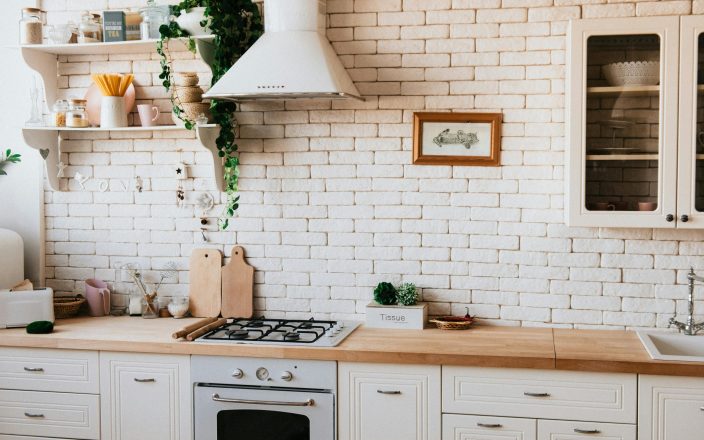 A kitchen with white brick walls, white cabinets and a wooden counter. There's light coming in through a window out of frame, and shelves with knickknacks on the walls.