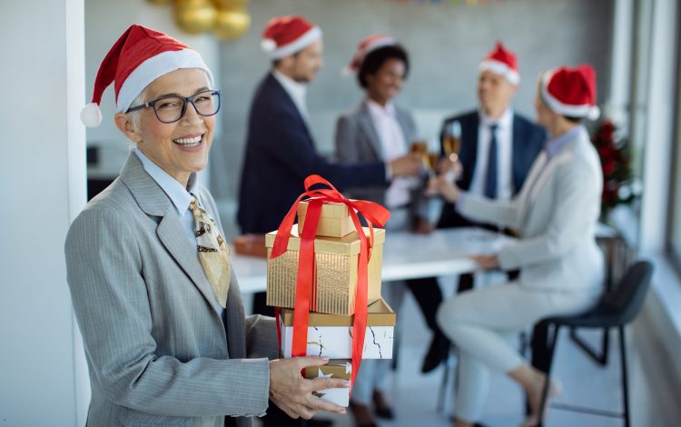 Happy mature businesswoman with gift boxes coming to Christmas party in the office and looking at camera. Her colleagues are celebrating in the background.