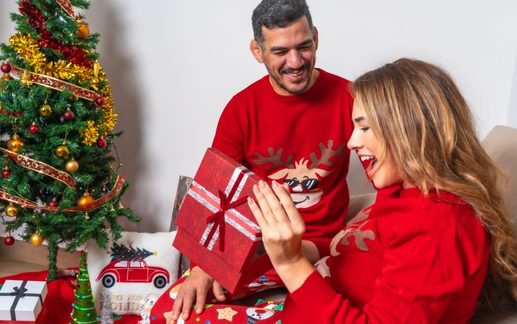 Young couple sitting on the sofa with decoration and red Christmas clothes, boyfriend giving a gift to the girlfriend and very happy girlfriend